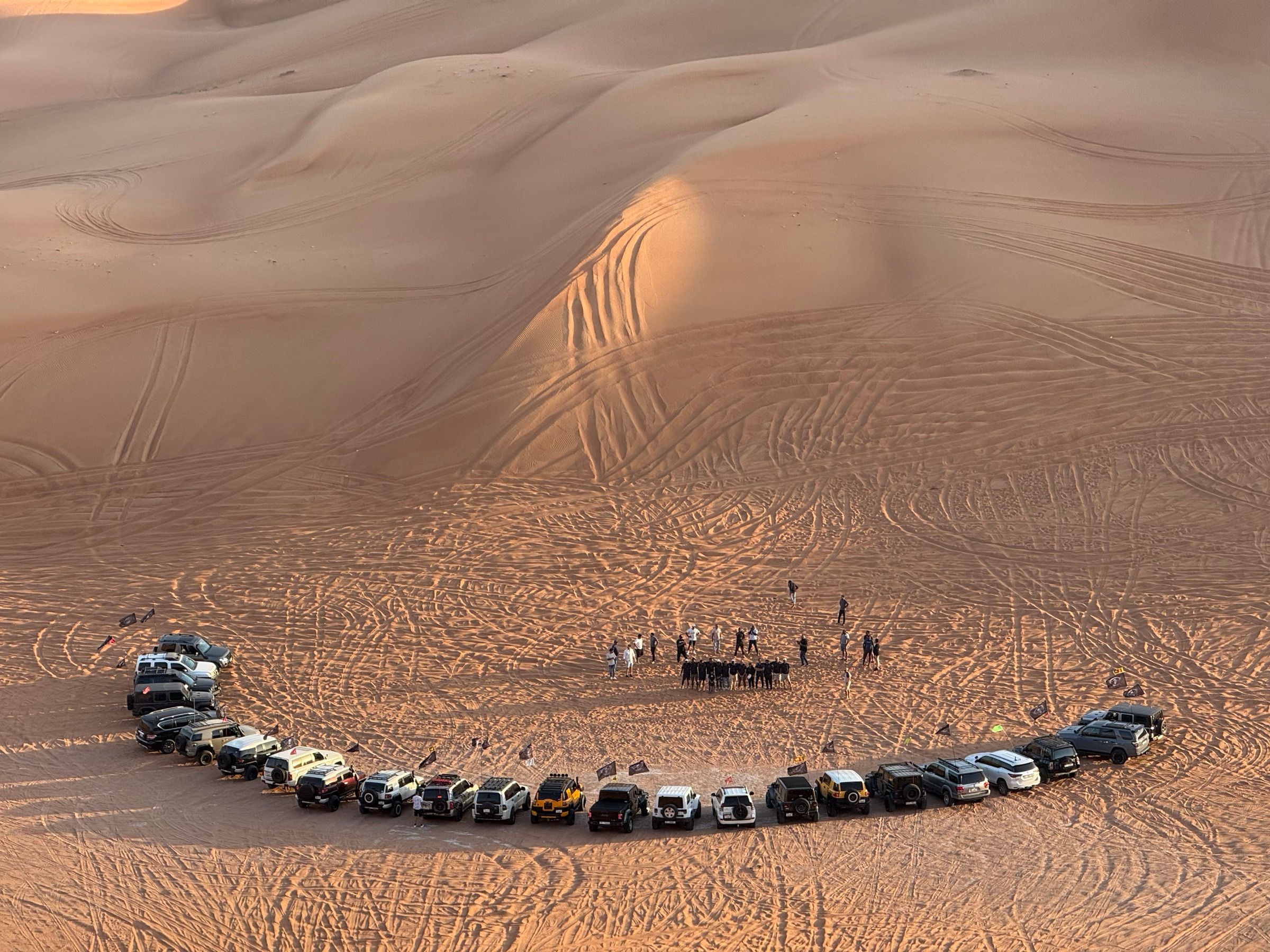 A convoy of 4x4s parked in a semicircle on the dunes at sunset while the group gathers for a briefing