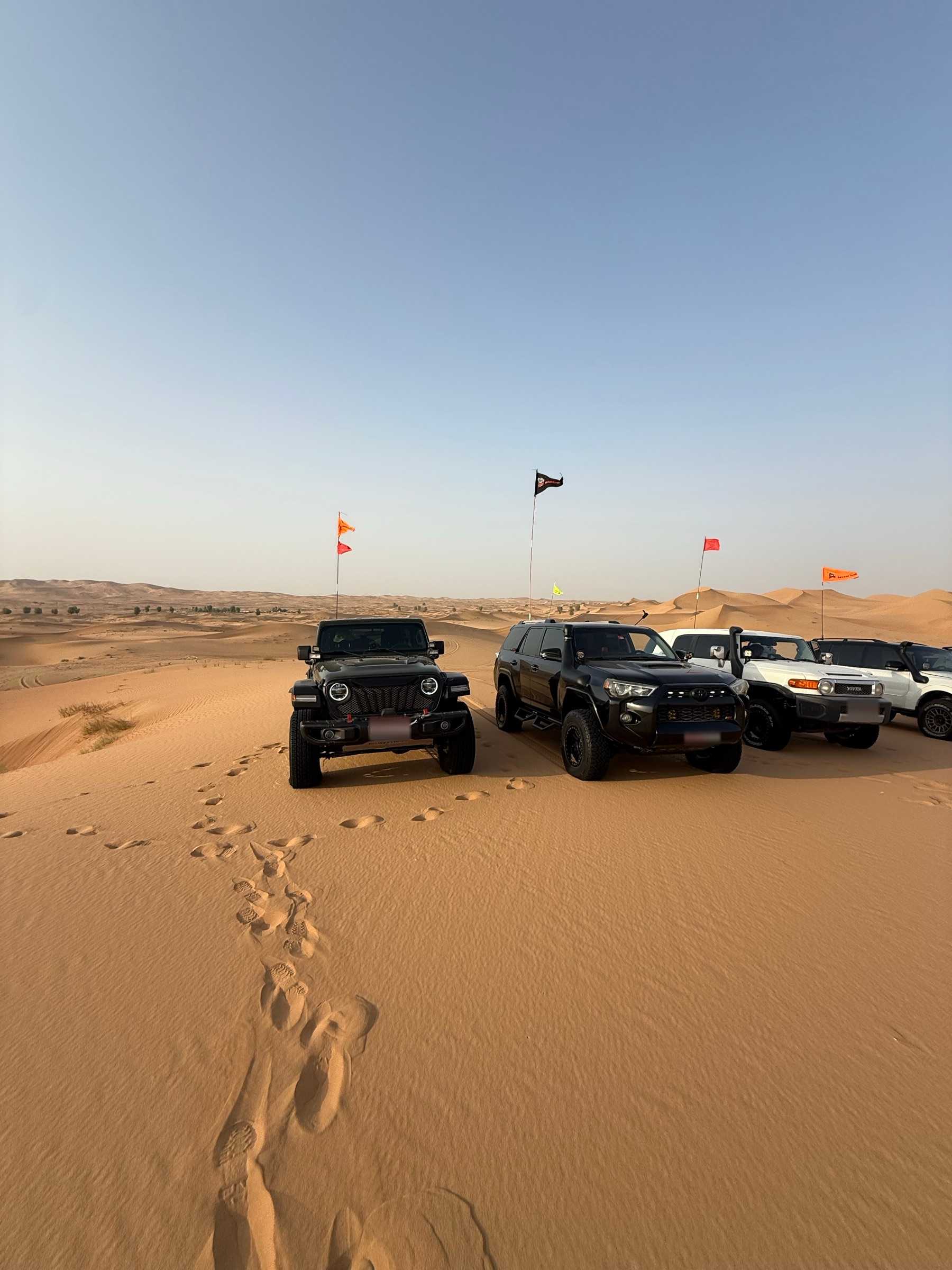 Four 4x4s parked side by side on sand among the dunes, with convoy flags on tall poles